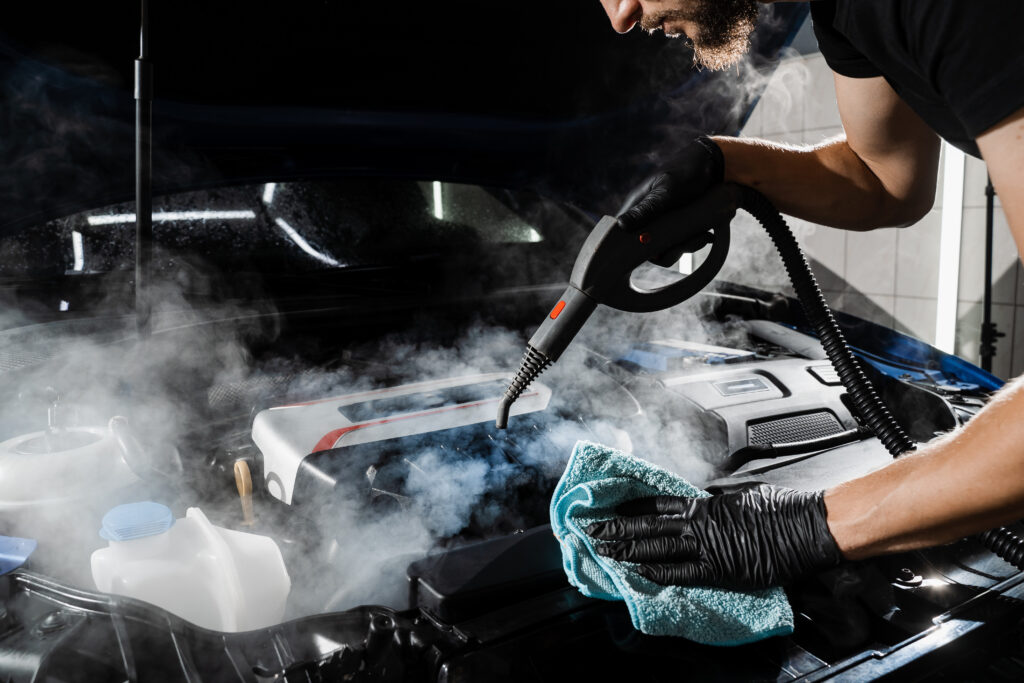 Technician steam cleaning and drying a car engine bay during a professional detailing service.