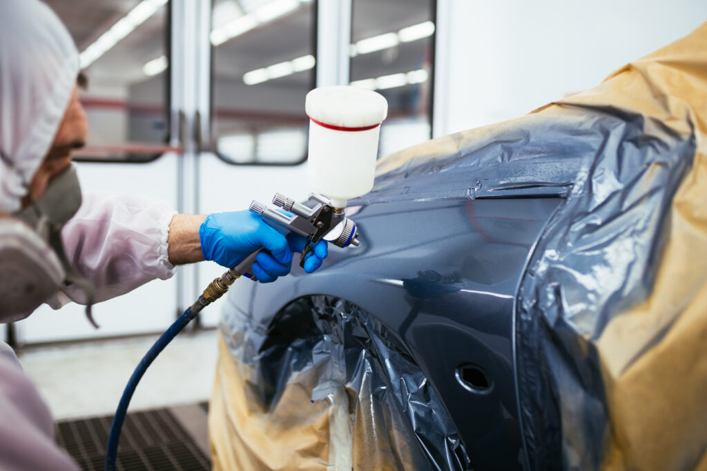 A technician spray painting a masked car panel inside an automotive paint booth.