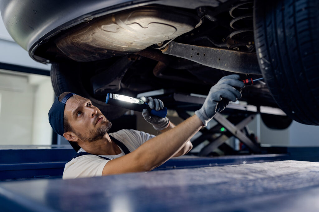 Mechanic inspecting the undercarriage of a car on a hoist using a flashlight during a repair shop inspection.